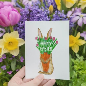 May include: A white card with a watercolor illustration of a brown bunny holding a bunch of carrots. The bunny is surrounded by green leaves and pink flowers. The text "Happy Easter" is written in white letters on the card.