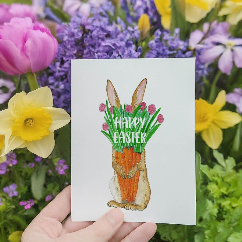 May include: A white card with a watercolor illustration of a brown bunny holding a bunch of carrots. The bunny is surrounded by green leaves and pink flowers. The text "Happy Easter" is written in white letters on the card.