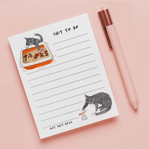 May include: A white notepad with horizontal lines and the text "SHIT TO DO" and "GET SHIT DONE". The notepad features illustrations of gray cats and a cat litter box. A pink pen with a rose gold clip rests on the right.