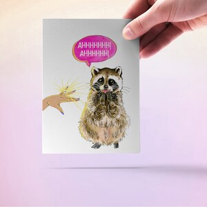 May include: A greeting card with a cartoon raccoon with a pink speech bubble that says "AHHHHHHH! AHHHHHHH!" The raccoon is looking up at a hand with a ring on it.