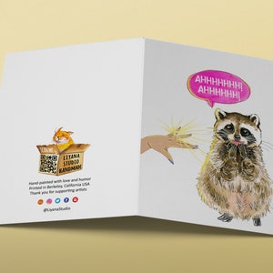 May include: A greeting card with a raccoon illustration and the text "AHHHHHHH! AHHHHHH!" in a pink speech bubble. The raccoon is looking up with a surprised expression and has its paws together. The card is white with a light yellow background.