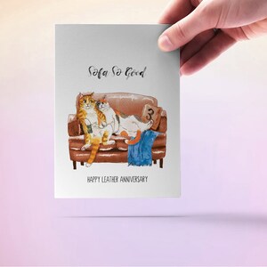 May include: A greeting card with a watercolor illustration of two cats sitting on a brown leather sofa. The text on the card reads "Sofa So Good" and "Happy Leather Anniversary".
