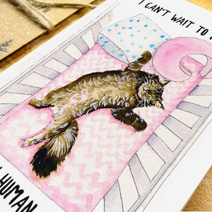 Op de afbeelding: Een aquarel-illustratie van een bruine tabby-kat die in een wieg ligt met een roze deken. De tekst "Ik kan niet wachten om mijn nieuwe mens te ontmoeten" staat op de kaart.