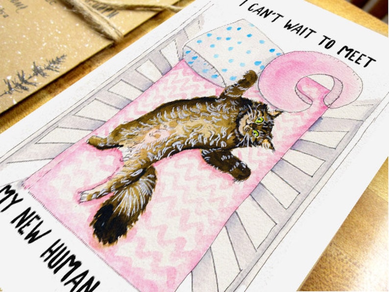 Op de afbeelding: Een aquarel-illustratie van een bruine tabby-kat die in een wieg ligt met een roze deken. De tekst "Ik kan niet wachten om mijn nieuwe mens te ontmoeten" staat op de kaart.
