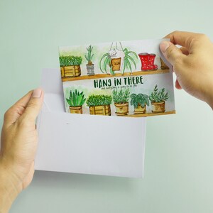 Peut inclure: Une carte de v&oelig;ux aquarelle avec une illustration de plantes en pot sur une &eacute;tag&egrave;re, en vert et marron. Le texte sur la carte est "Hang in there and everything is going to be fine."
