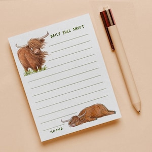 May include: A white notepad with green horizontal lines, featuring a brown Highland cow illustration at the top and another lying down at the bottom. The text reads DAILY BULL SHEET at the top and MOOOD at the bottom, accompanied by a multi-colored pen.