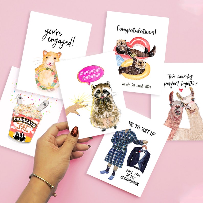 May include: Set of five greeting cards with humorous illustrations and text. The cards feature a raccoon, a hamster, two otters, two llamas, and a man in a plaid robe. The text on the cards includes "You're engaged!" "Congratulations!" "Two weirdos perfect together" "Me to suit up" and "Will you be my groomsman?"