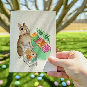 May include: A watercolor illustration of a brown rabbit pushing a shopping cart full of colorful Easter eggs. The cart is labeled "Easter Egg".