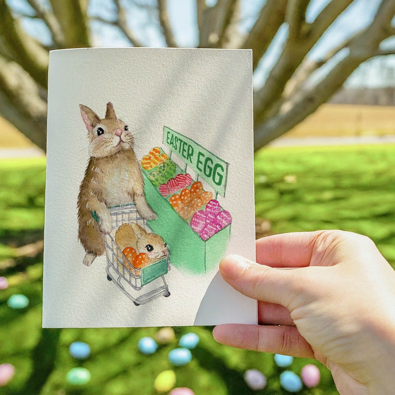 May include: A watercolor illustration of a brown rabbit pushing a shopping cart full of colorful Easter eggs. The cart is labeled "Easter Egg".