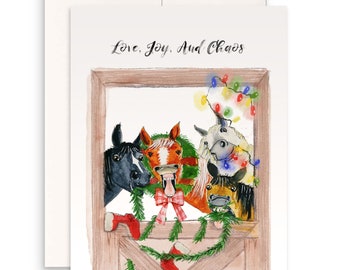 May include: A Christmas card featuring a watercolour illustration of four horses peeking out of a stable door. The horses are decorated with Christmas lights and greenery. The text "Love, Joy, And Chaos" is written at the top of the card.