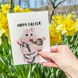 May include: A pink Easter bunny holding two kittens. The bunny is wearing a blue bow and the kittens are wearing bunny ears. The card says "Happy Easter".