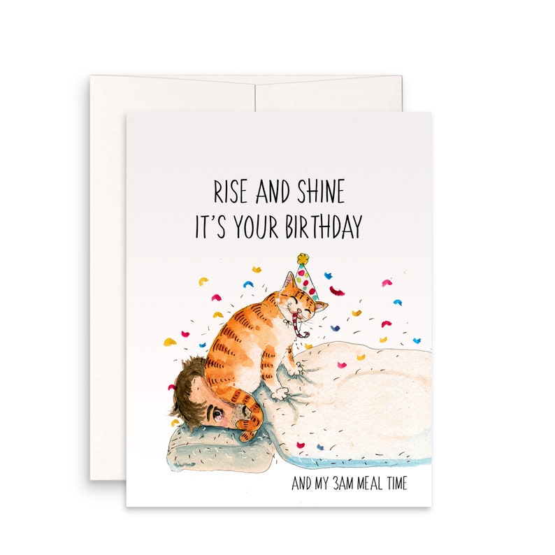 May include: A birthday card featuring a watercolour illustration of an orange tabby cat wearing a party hat and sitting on a person's head in bed. The card reads "Rise and Shine It's Your Birthday" and "And My 3AM Meal Time".