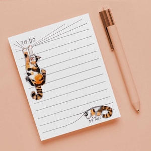 May include: A white notepad with horizontal lines and a cartoon cat illustration. The cat is orange, black, and white, hanging from the top of the page. The notepad has the words "TO DO" and "OR NOT". A pink pen with a gold clip rests beside the notepad.