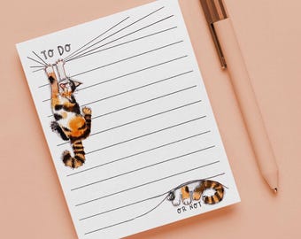 May include: A white notepad with horizontal lines and a cartoon cat illustration. The cat is orange, black, and white, hanging from the top of the page. The notepad has the words "TO DO" and "OR NOT". A pink pen with a gold clip rests beside the notepad.