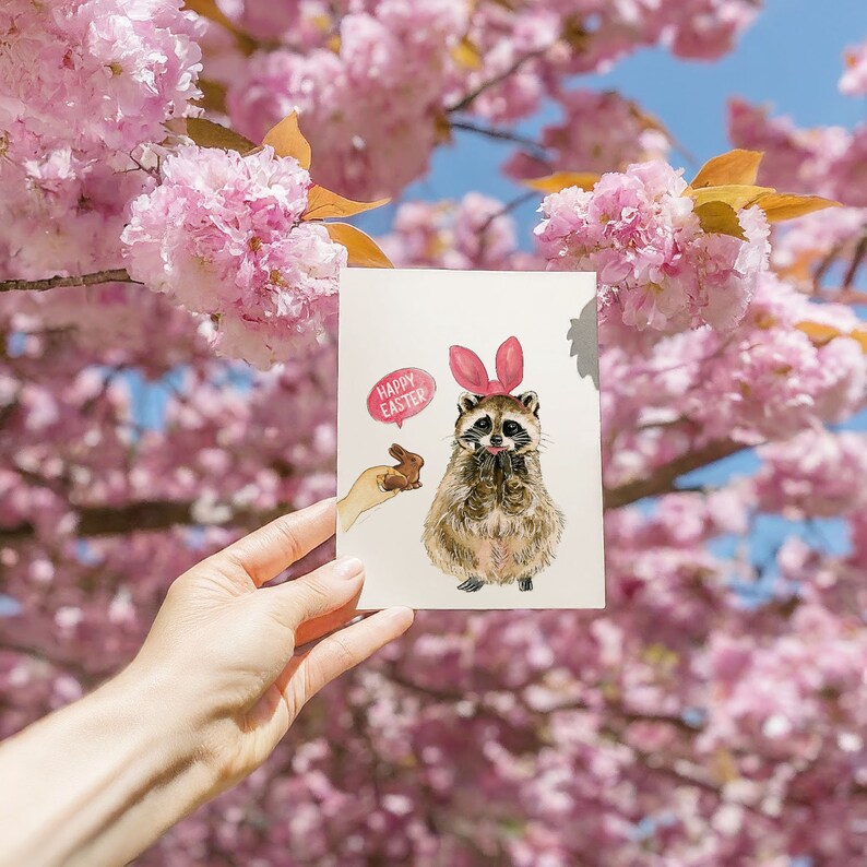 May include: A greeting card featuring a cartoon raccoon wearing a pink bunny ear headband and holding a chocolate bunny. The card has a pink speech bubble that says "Happy Easter".