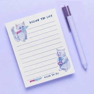 May include: A lined notepad with the text "KILLING THE LIST" and "KILLED 'EM ALL". The notepad features watercolor illustrations of gray cats holding a knife and scissors. A white pen with a silver clip rests on the side.