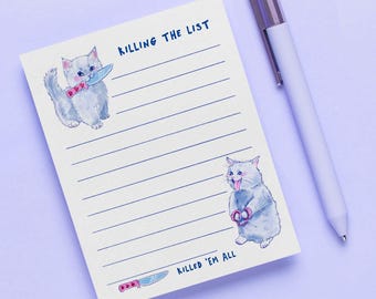 May include: A lined notepad with the text "KILLING THE LIST" and "KILLED 'EM ALL". The notepad features watercolor illustrations of gray cats holding a knife and scissors. A white pen with a silver clip rests on the side.
