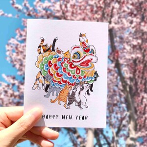 May include: A greeting card featuring a colorful illustration of a lion dance costume with multiple cats inside. The card has the text "HAPPY NEW YEAR" at the bottom. The background is blurred with pink and blue hues.