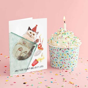 Pu&ograve; includere: Un biglietto di compleanno con un'illustrazione ad acquerello di un gatto con un cappello da festa, con la scritta "Another year? Knock it off!". Accanto, un cupcake in una tazza blu e bianca cosparsa di zuccherini, sormontato da glassa bianca e una candela accesa.