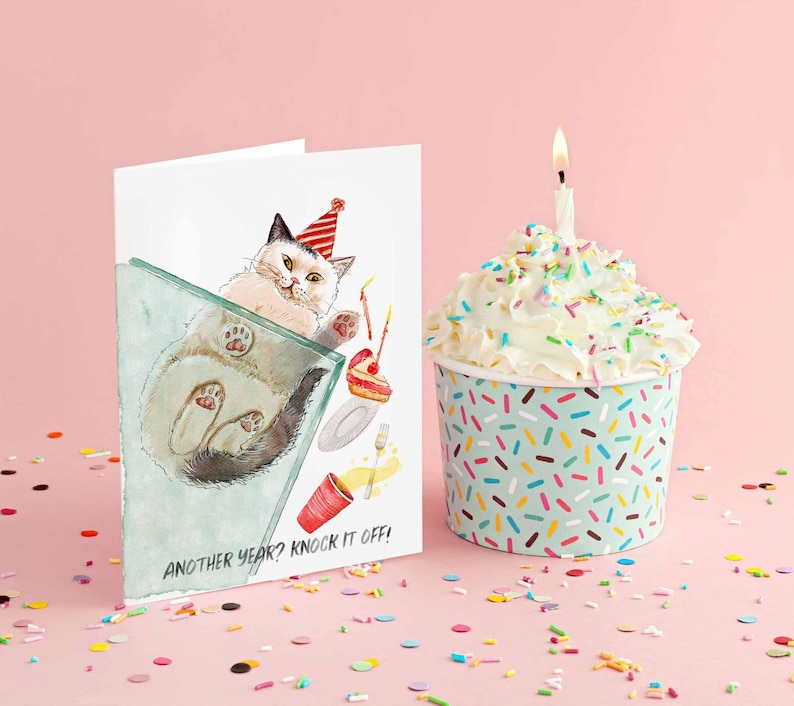 Pu&ograve; includere: Un biglietto di compleanno con un'illustrazione ad acquerello di un gatto con un cappello da festa, con la scritta "Another year? Knock it off!". Accanto, un cupcake in una tazza blu e bianca cosparsa di zuccherini, sormontato da glassa bianca e una candela accesa.