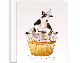 Op de afbeelding: Wenskaart met een aquarel illustratie van een koe met een feesthoedje, zittend op een cupcake met hagelslag. De kaart is van binnen leeg en bevat een witte envelop. De afbeelding staat op een witte achtergrond.