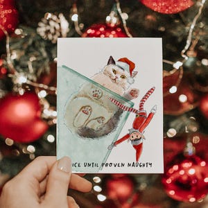 Pu&ograve; includere: Biglietto d'auguri natalizio con un'illustrazione ad acquerello di un gatto con un cappello da Babbo Natale e un elfo. La carta recita "NICE UNTIL PROVEN NAUGHTY". Lo sfondo presenta decorazioni natalizie.