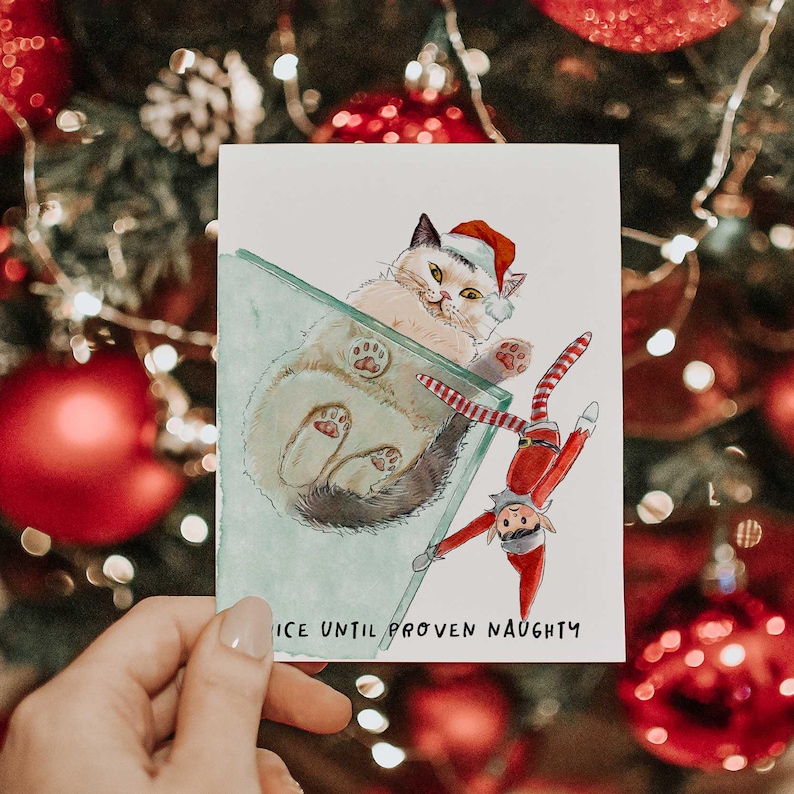 Pu&ograve; includere: Biglietto d'auguri natalizio con un'illustrazione ad acquerello di un gatto con un cappello da Babbo Natale e un elfo. La carta recita "NICE UNTIL PROVEN NAUGHTY". Lo sfondo presenta decorazioni natalizie.