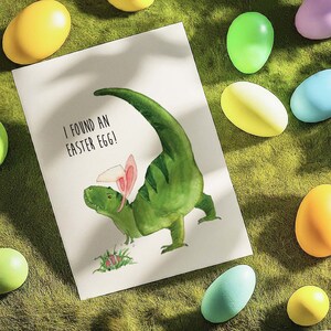 May include: A green dinosaur with pink bunny ears holds an Easter egg. The dinosaur is on a white card with the text "I found an Easter egg!"