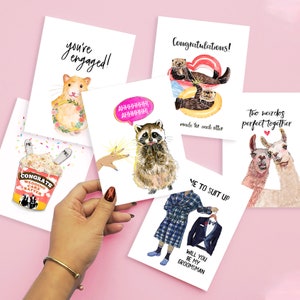 May include: Set of five greeting cards with humorous illustrations and text. The cards feature a raccoon, a hamster, two otters, two llamas, and a man in a plaid robe. The text on the cards includes "You're engaged!" "Congratulations!" "Two weirdos perfect together" "Me to suit up" and "Will you be my groomsman?"
