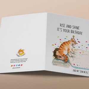 May include: A greeting card featuring a watercolour illustration of a cat wearing a party hat, waking someone up. The text on the card reads "Rise and shine it's your birthday and my 3am meal time."
