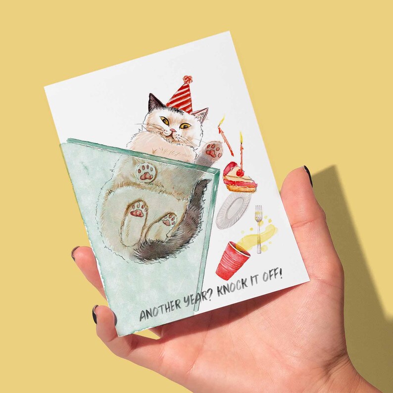 Pu&ograve; includere: Un biglietto di compleanno tenuto in mano presenta un'illustrazione ad acquerello di un gatto con un cappello da festa, all'interno di un bicchiere. La carta include una fetta di torta, una tazza rossa e il testo "ANOTHER YEAR? KNOCK IT OFF!"