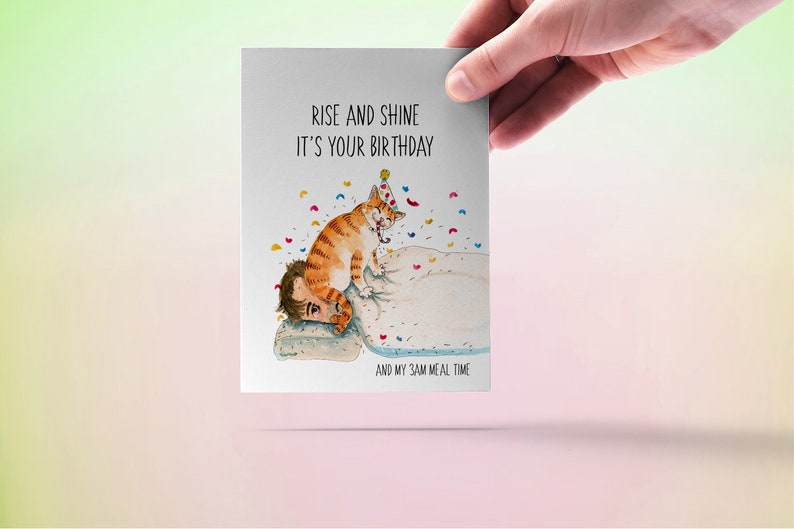 May include: A greeting card featuring a cartoon illustration of a cat wearing a party hat and waking someone up. The text on the card reads "Rise and shine it's your birthday and my 3am meal time."