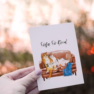 May include: A watercolor illustration of two cats relaxing on a brown couch. The text "Sofa So Good" is written in black cursive above the cats.