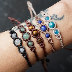 Könnte beinhalten: Fünf Makramee-Armbänder mit runden Edelsteinperlen in Weiß, Hellgrün, braunem Tigerauge, Dunkelblau und Türkis. Jedes Armband ist mit geknoteten Schnüren in Schwarz, Dunkelbraun, Mittelbraun, Beige und Creme gefertigt.