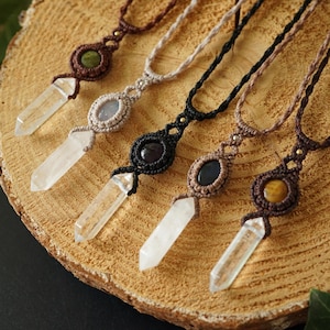 Pode incluir: Cinco colares de macramé feitos à mão com pingentes de cristal e detalhes de pedras preciosas. Os colares apresentam várias cores de fio de macramé e diferentes pedras preciosas, incluindo quartzo transparente, ônix preto e olho de tigre. Os pingentes são exibidos em uma superfície de madeira.
