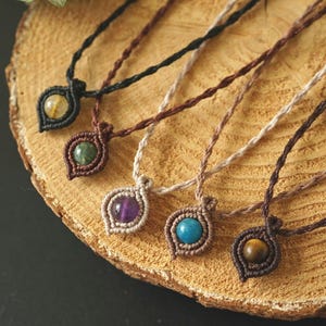 Può includere: Primo piano di sei collane in macramè con ciondoli di pietre preziose. I ciondoli presentano pietre di vari colori, tra cui giallo, verde, viola, blu e marrone. Le collane sono esposte su una superficie di legno.