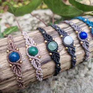 May include: A collection of macrame bracelets with various gemstone centerpieces, including tiger's eye, malachite, and amethyst. The bracelets are in shades of brown, black, and blue, with gold-colored bead accents. The bracelets are displayed on a piece of wood.