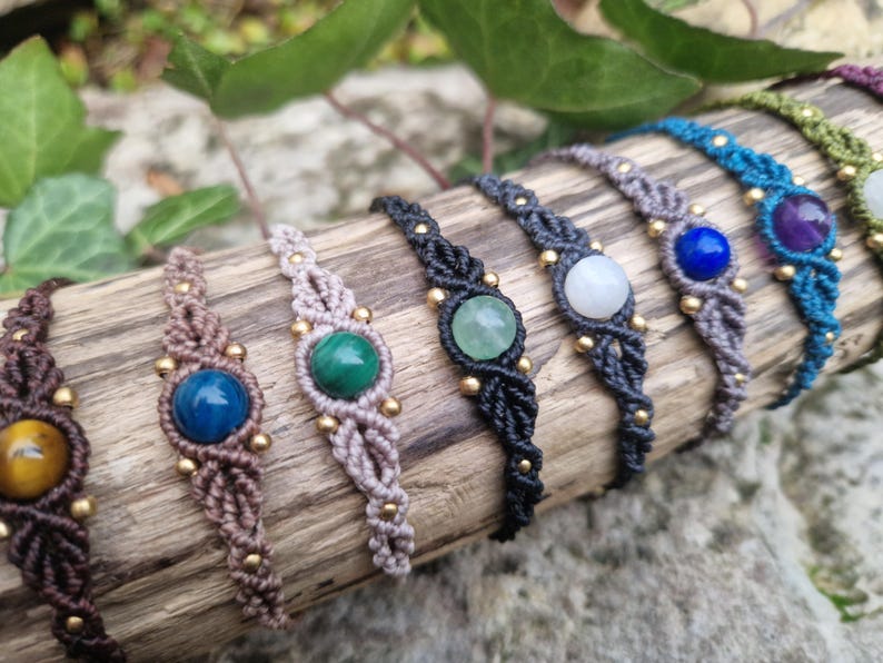 May include: A collection of macrame bracelets with various gemstone centerpieces, including tiger's eye, malachite, and amethyst. The bracelets are in shades of brown, black, and blue, with gold-colored bead accents. The bracelets are displayed on a piece of wood.