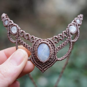 May include: A handcrafted macrame necklace featuring a central oval moonstone and smaller moonstones along the top. The necklace is made with brown cord and is held in a hand against a blurred background.