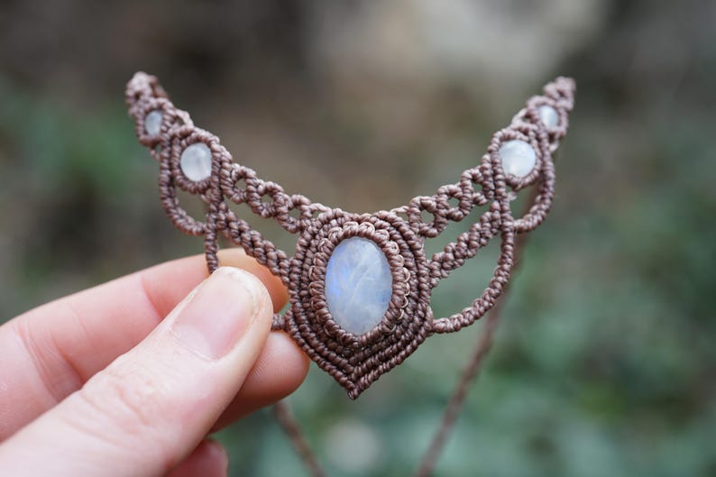 May include: A handcrafted macrame necklace featuring a central oval moonstone and smaller moonstones along the top. The necklace is made with brown cord and is held in a hand against a blurred background.