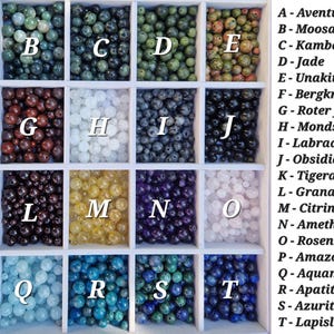 May include: A display of various gemstone beads in a white divided tray. The beads are arranged in rows and columns, each section labeled with a letter and a gemstone name. Colors include green, brown, white, black, yellow, purple, and blue.