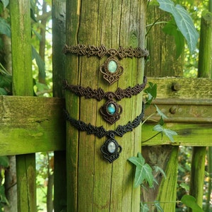 May include: Three macrame necklaces with gemstone pendants are displayed on a weathered wooden post. The necklaces are brown and black, with oval-shaped gemstones in shades of blue, green, and white. The background features a green wooden fence and foliage.