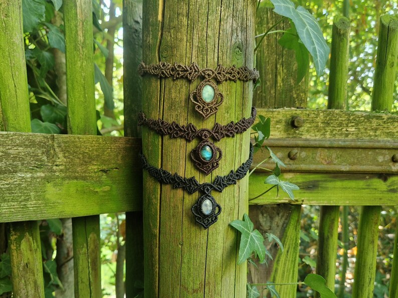 May include: Three macrame necklaces with gemstone pendants are displayed on a weathered wooden post. The necklaces are brown and black, with oval-shaped gemstones in shades of blue, green, and white. The background features a green wooden fence and foliage.
