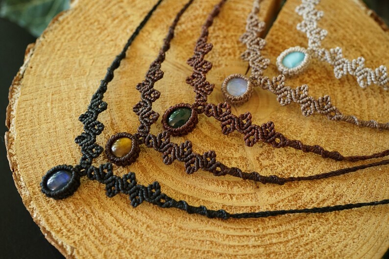 Puede incluir: Cuatro collares con intrincados cordones de macram&eacute; en negro, marr&oacute;n oscuro, marr&oacute;n medio y beige. Cada collar se centra en una sola piedra preciosa ovalada, incluyendo piedras azules, ojo de tigre, verde oscuro, azul claro y blanco iridiscente.