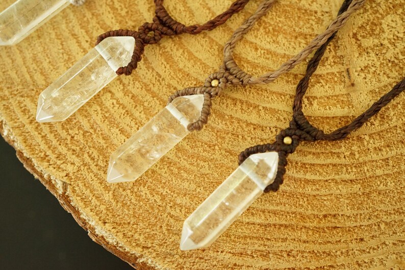 May include: Three necklaces featuring clear, faceted, hexagonal crystal points suspended from braided cord. Each pendant is secured with a macrame knot and a small, round, metallic bead. The cords vary in shades of brown, from light tan to dark brown.