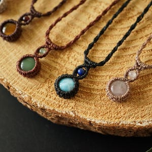May include: Close-up of several handmade macrame necklaces with gemstone pendants. The necklaces feature various colored stones, including green, blue, and pink, set in intricate macrame knots. The necklaces are displayed on a wooden surface.