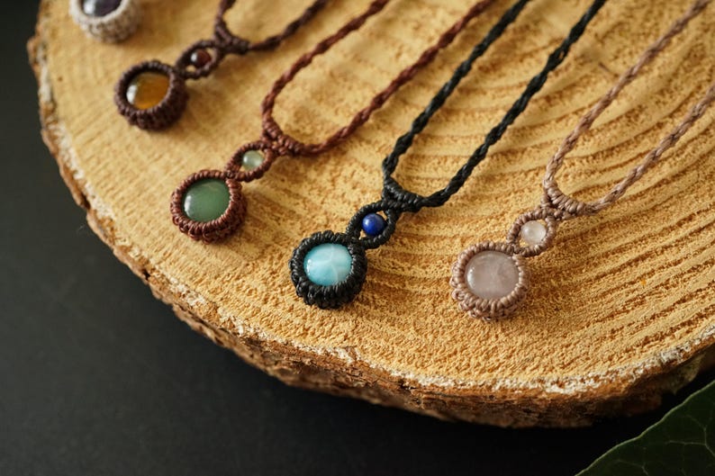 May include: Close-up of several handmade macrame necklaces with gemstone pendants. The necklaces feature various colored stones, including green, blue, and pink, set in intricate macrame knots. The necklaces are displayed on a wooden surface.