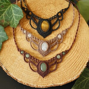May include: Four macrame necklaces with gemstone pendants are displayed on a wooden surface, surrounded by green leaves. The necklaces feature brown, tan, and black cords, with oval and round gemstones in shades of green, white, and yellow.