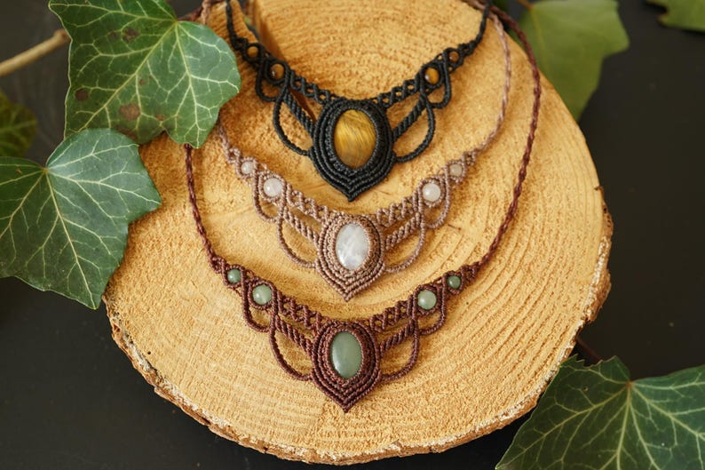 May include: Four macrame necklaces with gemstone pendants are displayed on a wooden surface, surrounded by green leaves. The necklaces feature brown, tan, and black cords, with oval and round gemstones in shades of green, white, and yellow.