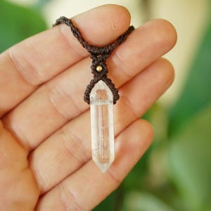 May include: A clear, double-terminated quartz crystal pendant secured in a dark brown macrame cord setting. The necklace features a small, round, metallic gold-colored bead accent positioned directly above the crystal point in the woven cord.